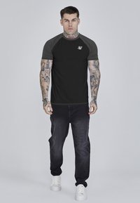 Schwarzes T-Shirt mit kurzen Ärmeln und grauen Raglanärmeln, ausgestattet mit einem kleinen weißen Logo auf der Brust, kombiniert mit dunklen Jeans und weißen Sneakers.