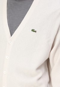 Lys gråstrikket cardigan med V-hals, fem knapper og en liten grønn krokodillelogo på venstre side. Strukturert stoff.