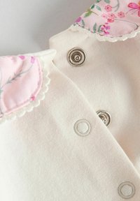 Chemise en tissu blanc avec un col floral rose, présentant des bords festonnés et des boutons-pression en argent pour la fermeture. Texture lisse dans l'ensemble.