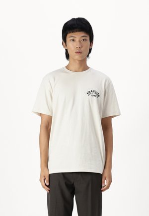 T-shirt beige in cotone con maniche corte, scollatura rotonda, caratterizzata da una stampa logo nera sul lato sinistro del petto. Abbinata a pantaloni scuri.