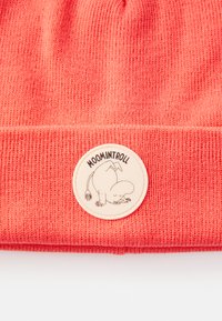 Koraļļu adīta beanie cepure ar maigu tekstūru, kas rotāta ar apaļu krēmkrāsas etiķeti, attēlojošu Moomintroll jautrā pozā.