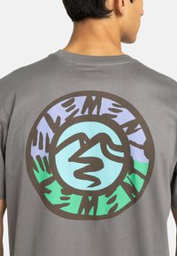 Grijze t-shirt met een cirkelvormige grafiek op de rug, in tinten van groen, blauw, paars en zwart, die abstracte bergen en landschapselementen weergeeft.