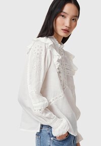 Blusa in cotone bianca con colletto e polsini arricciati, ricami intricati e dettagli in pizzo sulle maniche; vestibilità rilassata e texture leggermente trasparente.
