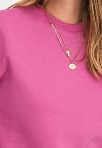 Sudadera de color rosa brillante con una textura suave y cuello redondo, combinada con dos collares dorados que tienen diferentes colgantes.