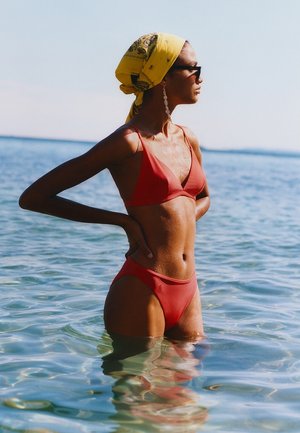 Kobieta w czerwonym bikini i żółtej chustce na głowie stoi do pasa w spokojnej wodzie morskiej, patrząc w prawo, ręce na biodrach.