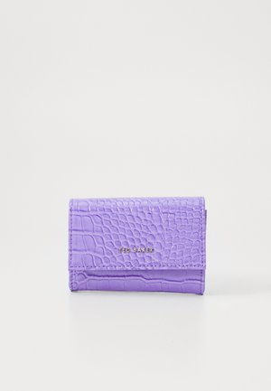 Lavandas violetā krāsā krokodila tekstūras ādas maks ar zelta "Ted Baker" logo uz priekšējās atloka, attēlots aizvērtā stāvoklī uz baltas fona.