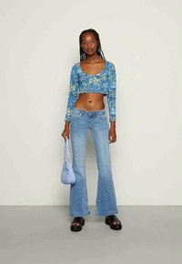 Modra cvetna kratka bluzica z dolgimi rokavi, svetlo modri široki kavbojke, črne platforme in svetlo modra torbica čez ramo. Gladke teksture, sproščen slog.