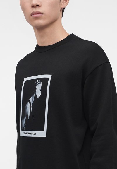 Homme portant un sweat-shirt noir avec un graphisme en noir et blanc d'une personne et le logo Emporio Armani à l'avant.