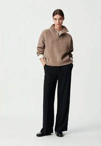 Khaki gerippter Strickpullover mit Reißverschlusskragen, über einem weißen Hemd getragen; kombiniert mit schwarzen, weit geschnittenen Hosen und schwarzen Schuhen.