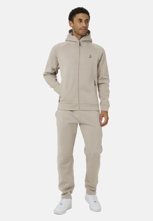 Hombre con chaqueta beige con capucha y cremallera y pantalones a juego con zapatillas blancas, de pie con una mano en el bolsillo mirando hacia la derecha.