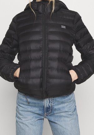 Personne portant une doudoune noire à capuche avec une fermeture éclair à l'avant et les mains dans les poches, associée à un jean bleu clair.