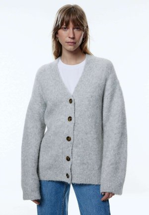 Hellgrauer Strickcardigan mit V-Ausschnitt, der vorne vier große braune Knöpfe hat und lockere, oversized Ärmel besitzt.