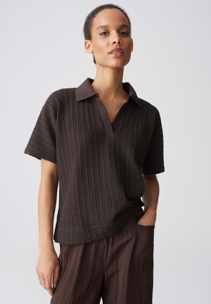 Frau trägt ein dunkelbraunes strukturiertes Kurzarmhemd mit Kragen und passende weite Hosen, steht mit einer Hand in der Tasche vor schlichtem Hintergrund.