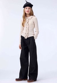 Világos bézs bordázott pulóver fodros elejjel, széles szárú fekete nadrággal és barna csizmával párosítva; a modell fekete barettet visel.