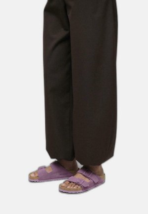 Lower legs wearing loose dark brown pants and purple Birkenstock sandals with two adjustable straps on bare feet.