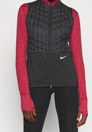 Personne portant une veste matelassée noire zippée sur un t-shirt rouge à manches longues et des leggings noirs avec un logo Nike blanc sur la veste.