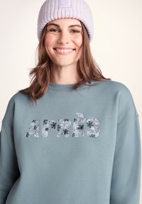Light blue sweatshirt with floral printed "APRÈS" text, ribbed crew neckline, and long sleeves. Paired with a light purple knit beanie.