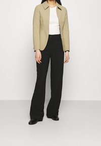 Veste beige à fermeture éclair sur un haut blanc, associée à un pantalon noir évasé. Tissu lisse, coupe structurée, et détail subtil au col. Chaussures noires.