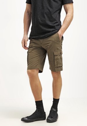 Mężczyzna ubrany w brązowe szorty cargo, czarny t-shirt z plamkami, czarne skarpetki i czarne trampki, stoi z jedną ręką w kieszeni.