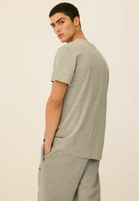 Szara bawełniana koszulka z krótkim rękawem, okrągłym dekoltem i luźnym krojem. Model stoi w pozycji obróconej, nosząc dopasowane szare dresy.