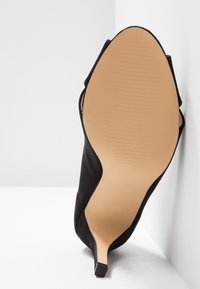 Černá nubuková stiletto bota na vysokém podpatku s hladkým béžovým podešví. Má špičatou špičku a nakloněné strany, což zdůrazňuje její elegantní tvar.