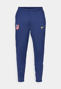 Marine atletiske bukser lavet af let stof, med et gult Nike-logo og et klubemblem med røde og hvide striber. Elastisk talje inkluderet.