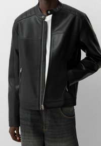 Giubbotto in ecopelle nera con colletto alto, apertura con zip e due tasche laterali. Presenta dettagli spalline trapuntate e una texture liscia.