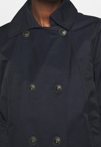 Abrigo de doble botonadura en azul marino oscuro con gran solapa y botones negros de cuatro agujeros, llevado por una persona que muestra el pecho superior y el cuello.