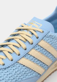 Zapatilla azul claro con material tejido texturizado, detalles de goma beige, y tres rayas beige. Presenta cordones planos beige y una marca dorada.