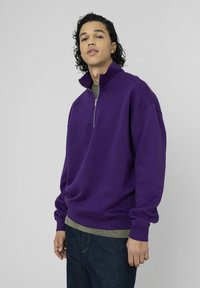 Sweat-shirt violet à fermeture éclair avec col montant, coupe décontractée et poignets côtelés. Porté par-dessus une chemise verte, associé à un jean en denim bleu.
