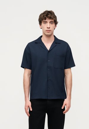 Junger Mann mit lockigem Haar, der ein kurzärmeliges marineblaues Hemd mit Knöpfen und schwarze Hose trägt, steht vor einem einfarbigen hellen Hintergrund.