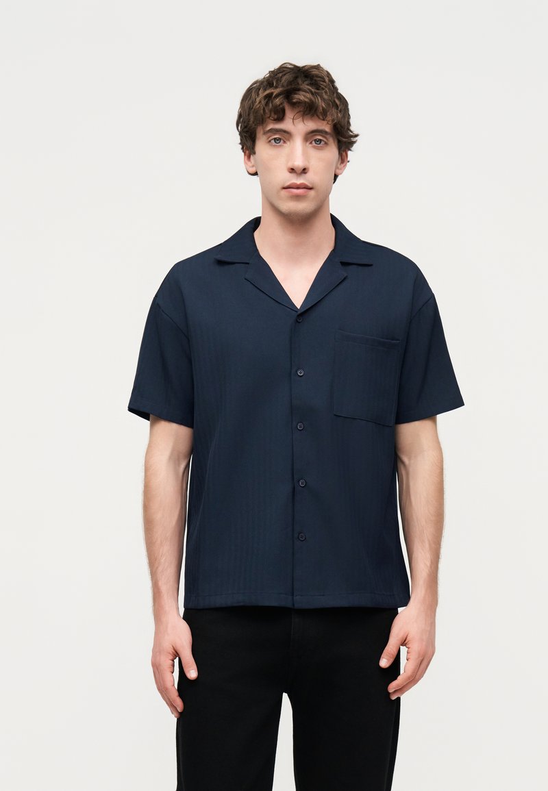 Νέος άνδρας με σγουρά μαλλιά που φορά κοντομάνικο μπλε σκούρο πουκάμισο με κουμπιά και μαύρο παντελόνι, στέκεται μπροστά σε ένα απλό φωτεινό φόντο.