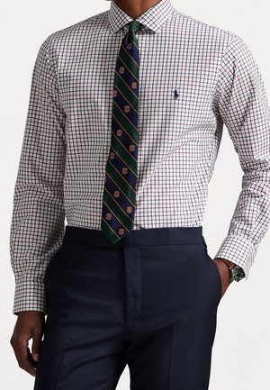 Homme portant une chemise blanche à carreaux, une cravate rayée marine et verte, un pantalon marine et une montre-bracelet, main dans la poche.