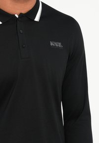Crna polo majica dugih rukava s bijelim prugastim ovratnikom, koja ima crno izvezeno "KARL LAGERFELD" logotip na lijevoj strani prsnog područja.