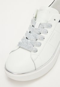 Valkoinen nahkainen sneaker, jossa on pyöreä kärki, harmaat nauhat ja minimaalinen brändäys. Ominaisuuksiin kuuluu sileä pinta ja paksu valkoinen pohja hopeisilla yksityiskohdilla.