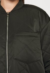 Kurtka bomberka pikowana w ciemnozielonym materiale z prążkowanym kołnierzem i srebrnym zamkiem błyskawicznym, wyposażona w kieszeń na piersi i wyraźny wzór diamentowy.