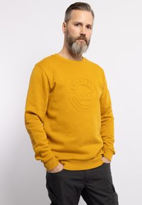 Sudadera amarilla con cuello redondo, detalle de logo texturizado en la parte frontal, puños acanalados y un corte relajado, combinada con pantalones oscuros.