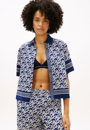 Femme aux cheveux bouclés portant une chemise à manches courtes à motifs bleu marine et blanc avec un pantalon assorti, avec un bralette noir Tommy Hilfiger visible.