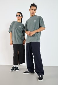Grå oversized t-shirts med logo foran, sort denimnederdel med sideskæring, og sorte sneakers med hvide accenter. Tilbehør inkluderer hvide solbriller.