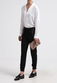 Hvid langærmet bluse med V-hals, sorte tapered bukser med lynlåse, sorte sko, holder en beige clutch. Glat stof, rene linjer.