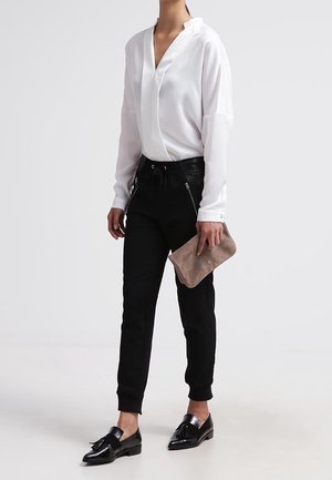 Biała bluzka z długim rękawem i dekoltem w kształcie litery V, czarne cygaretki z zamkami, czarne buty, trzymająca beżową kopertówkę. Gładki materiał, czyste linie.