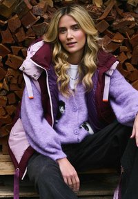 Fioletowy puszysty kardigan z czarno-białymi wzorami, noszony pod bordową i jasnoróżową kamizelką. Ciemne dżinsy i prosty biały t-shirt.