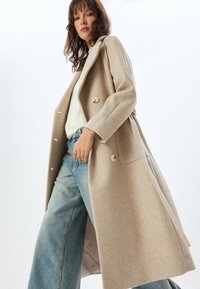 Abrigo de lana beige con un diseño entallado, solapas en V, dos botones, bolsillos laterales y una textura suave, combinado con jeans de pierna ancha azul claro.