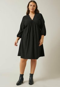 Schwarzes, knielanges Kleid mit tiefem V-Ausschnitt, unter der Brust gerafft und dreiviertellangen Puffärmeln. Getragen mit schwarzen Stiefeletten.