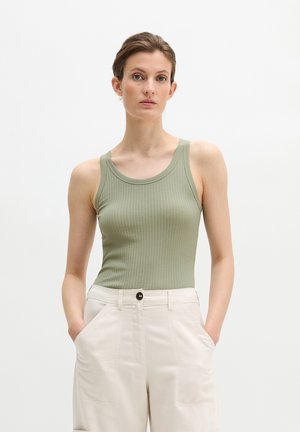 Vrouw met een lichtgroen geribd mouwloos topje en witte, hooggetailleerde broek met handen in de zakken, tegen een effen witte achtergrond.