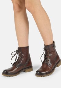 Bottines en cuir marron à lacets avec une semelle robuste, présentant une finition lisse, des accents cousus et un détail de sangle décoratif.