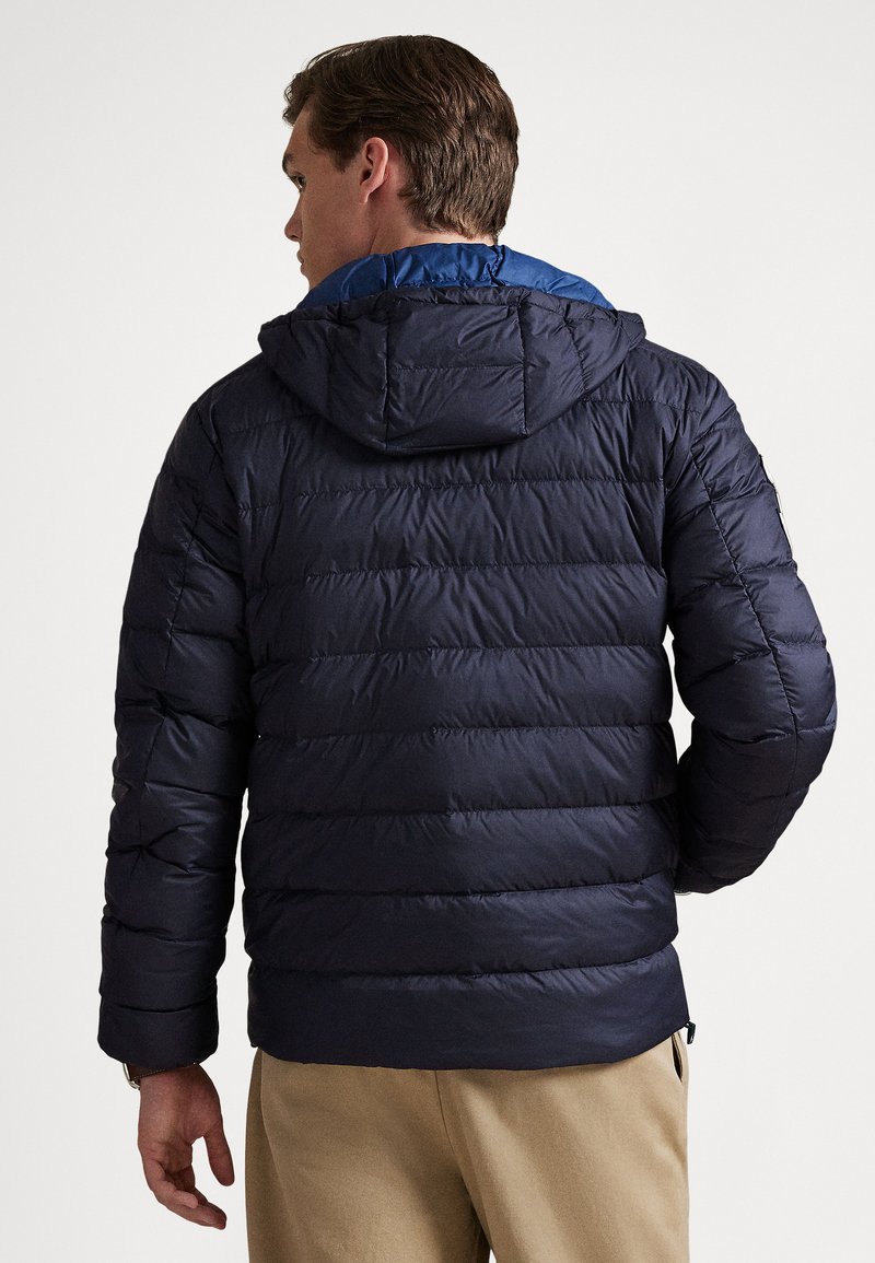 Man wearing a navy blue quilted puffer jacket with a hood and beige pants, facing away against a plain light background.