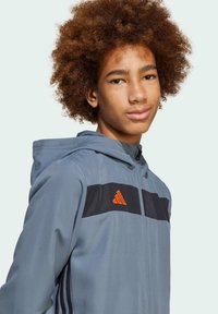 Veste de sport grise avec capuche, bande noire sur la poitrine et logo Adidas orange. Tissu lisse et détail de fermeture éclair à l'avant.