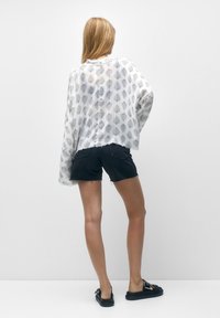 Biała przezroczysta bluzka z czarnymi wzorami liści, luźny krój, w połączeniu z czarnymi szortami dżinsowymi i czarnymi sandałami, stojąca na jednolitym tle.