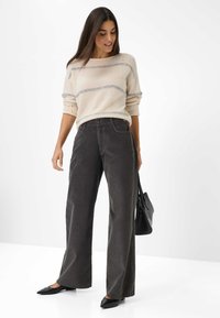 Helle beiges Sweatshirt mit Streifen, kombiniert mit dunkelgrauen weit geschnittenen Hosen. Modell hält eine schwarze Handtasche und trägt spitze schwarze Schuhe.